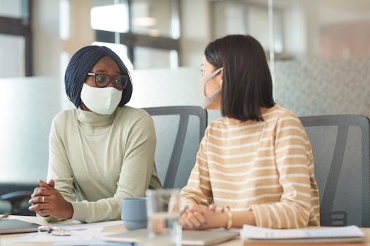 Two women wearing masks discuss work at an office table.