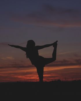 Silhouette of a woman practicing yoga at sunset, balancing on one leg in a peaceful outdoor setting.