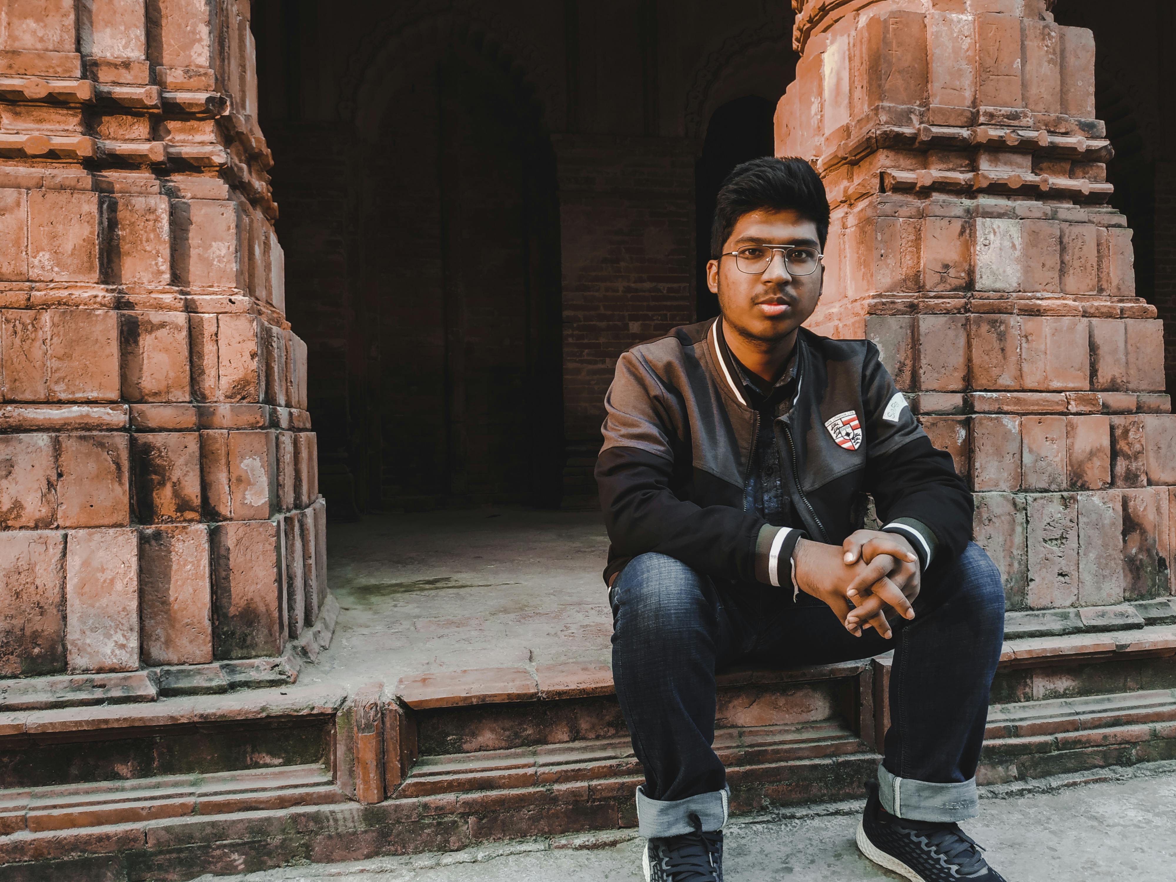 A young man in a black jacket and eyeglasses sitting on stone steps of a historical building.