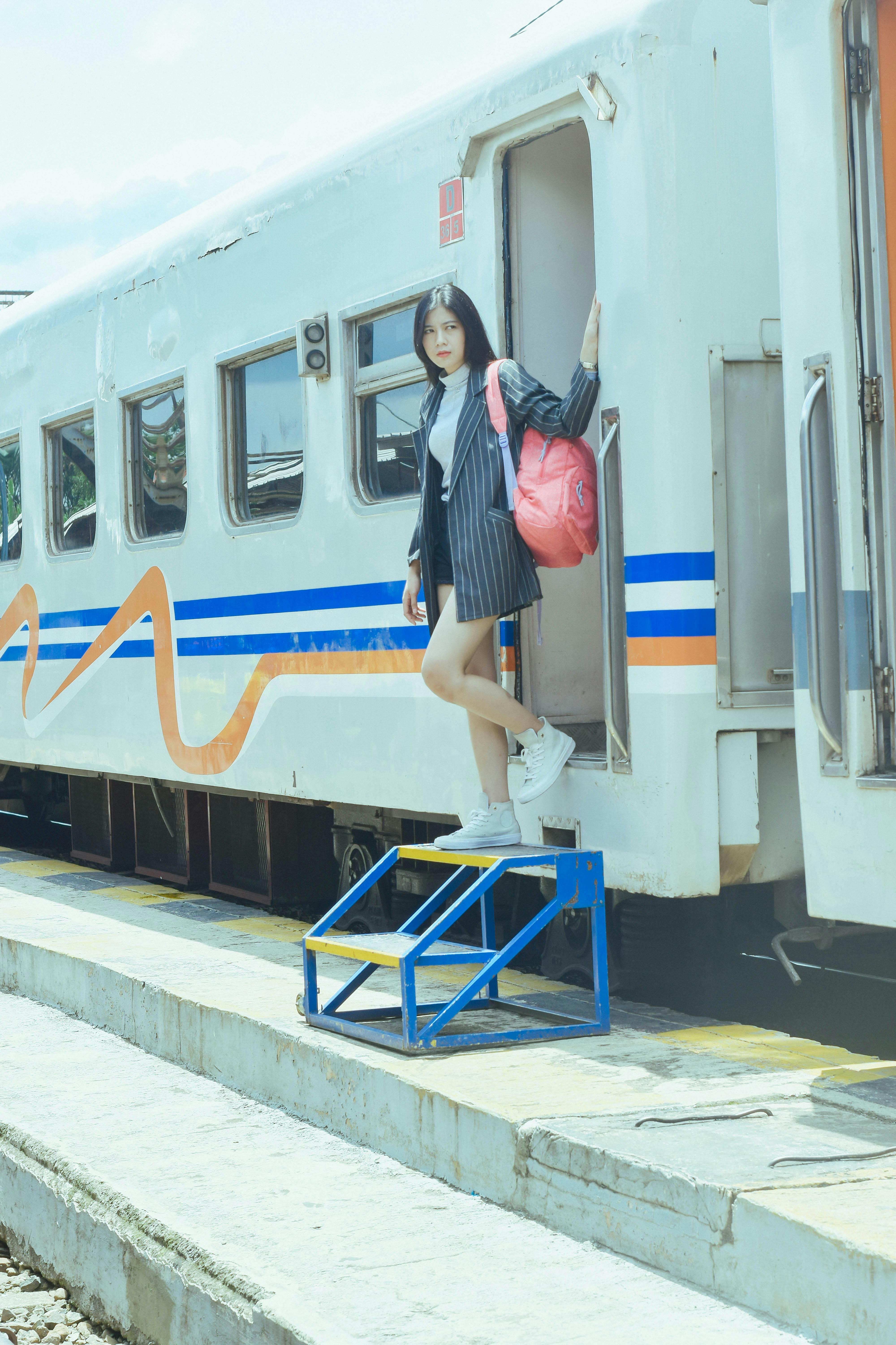 Stylish young woman with red backpack posing by train at West Java station.