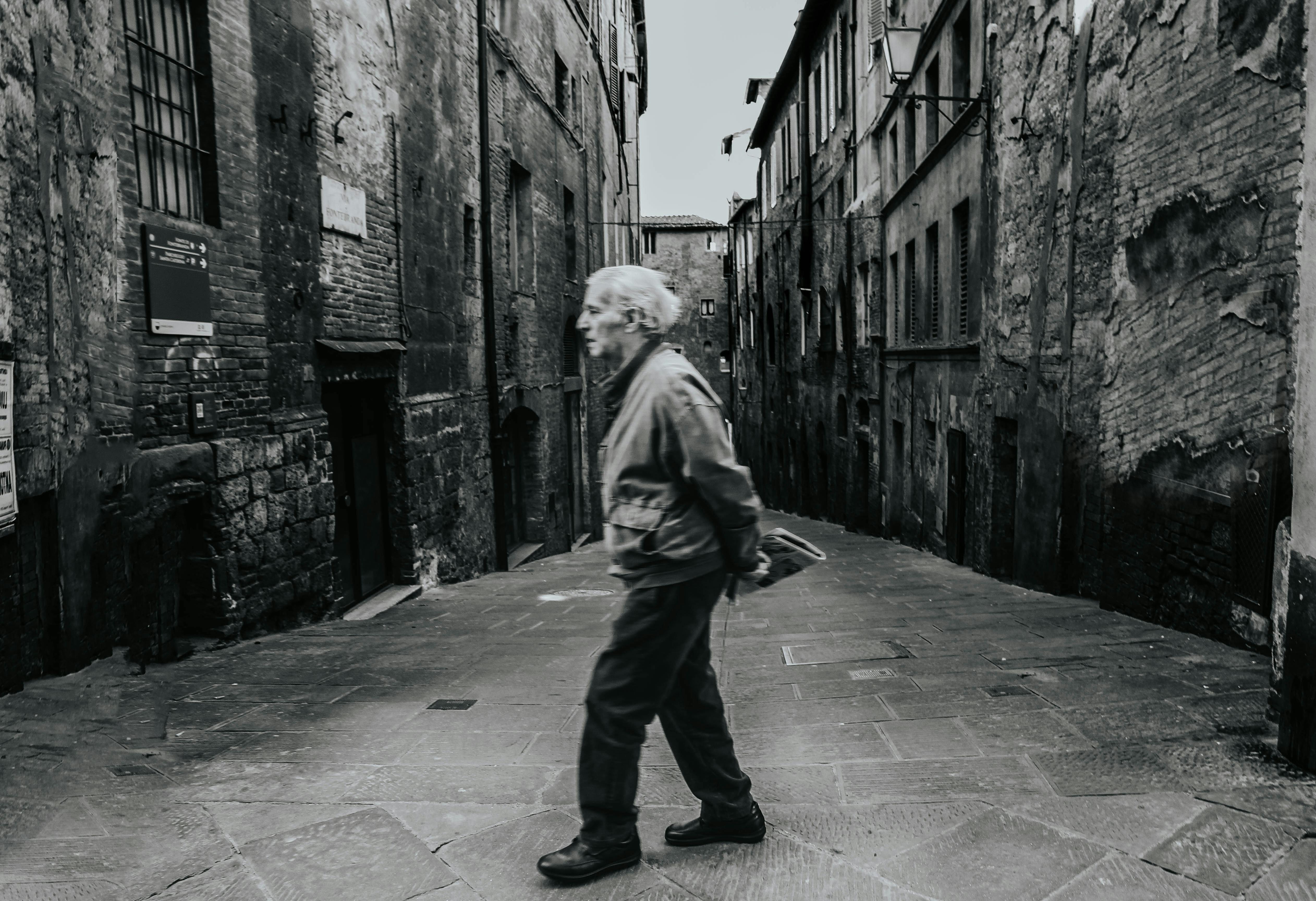 A senior man walking through a narrow, historic alley in Italy. Grayscale imagery.