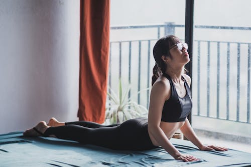 Full body of active barefoot Asian female in sportswear and eyeglasses exercising High cobra asana while practicing yoga at home