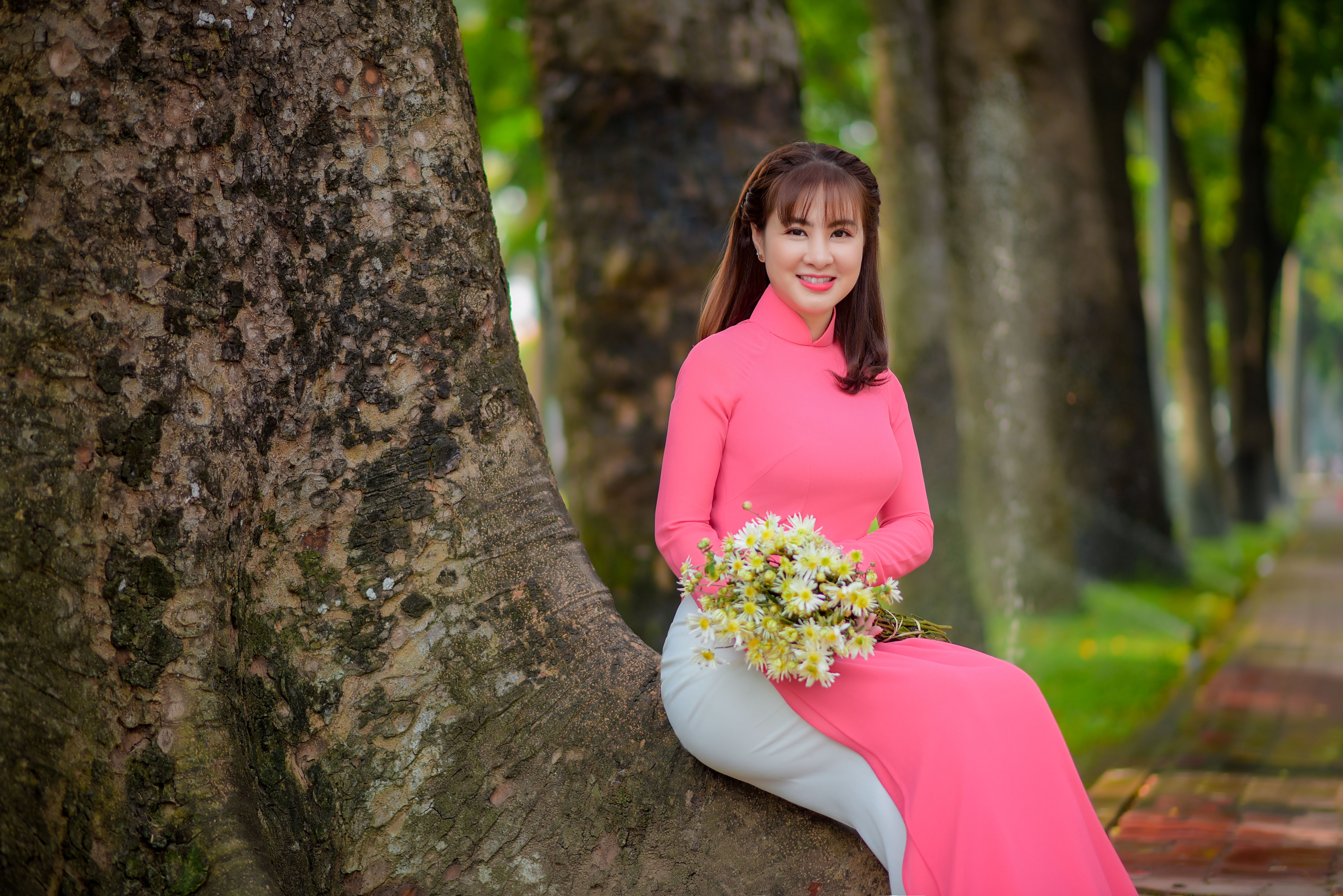 A graceful Vietnamese woman in a pink ao dai, sitting outdoors with a bouquet, exuding beauty and charm.