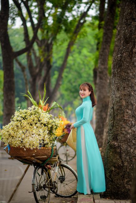 Elegant woman in turquoise ao dai with bicycle and flowers on a tree-lined street.