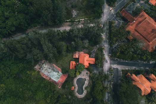 Aerial view of various residential houses with red roofs located in lush tropical forest near small pond and sports ground