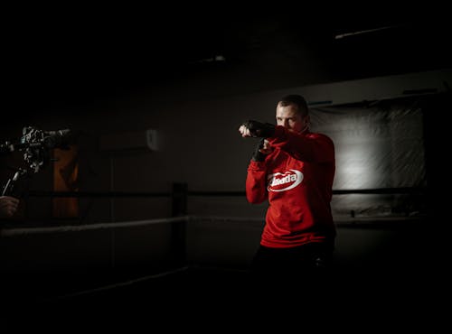 Powerful male athlete in sportswear practicing boxing hits in ring against crop anonymous partner recording video on photo camera