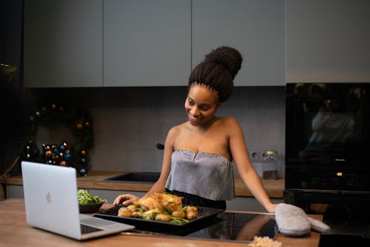African American woman in kitchen smiling at a laptop with a tray of roast chicken and vegetables.