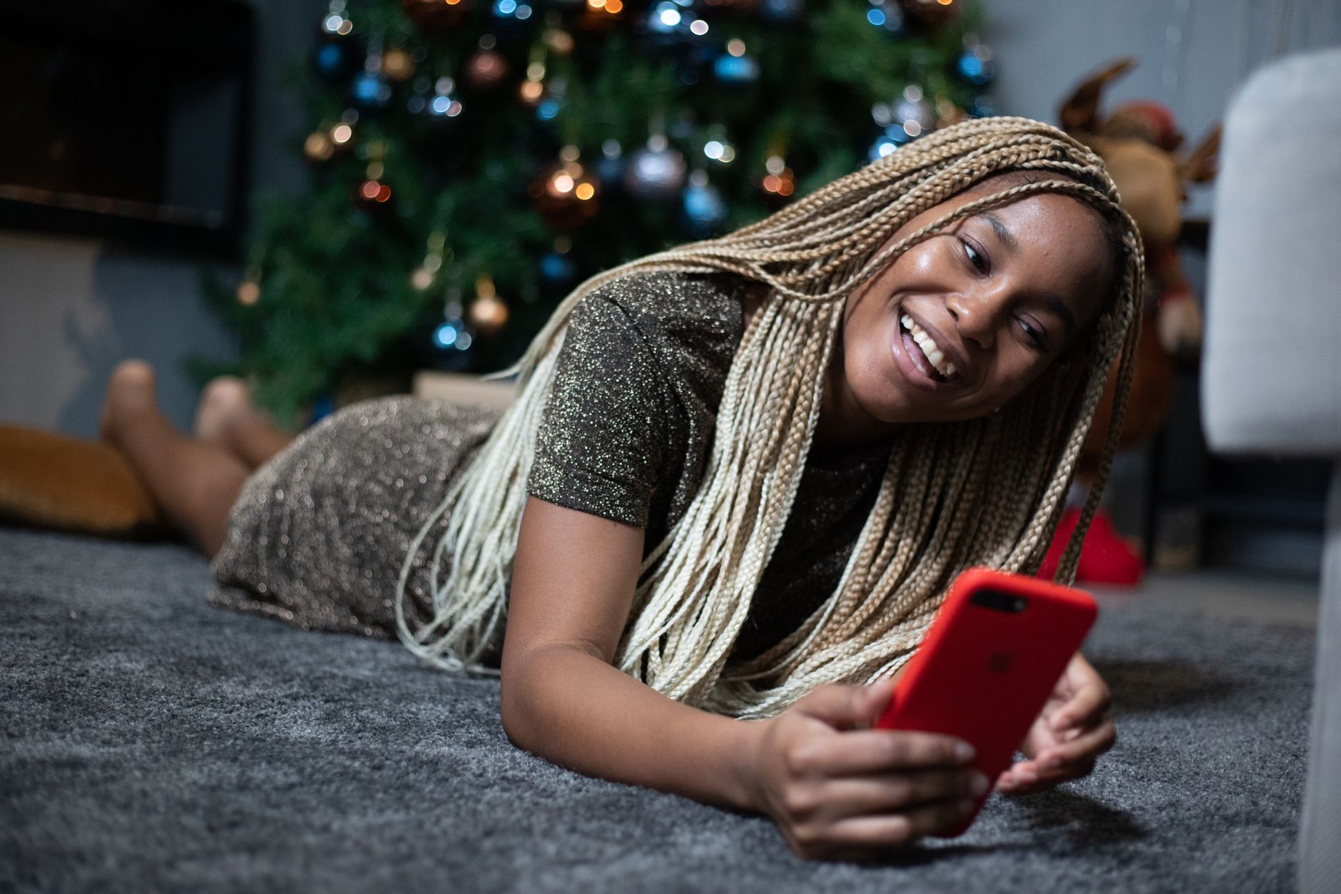 Giovane donna sorridente sdraiata su un tappeto, smartphone rosso in mano, davanti a un albero di Natale illuminato