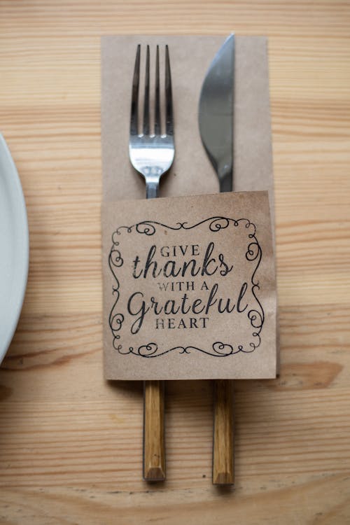 Top view of metal fork and knife in paper cover decorated with Give Thanks With A Grateful Heart inscription placed on wooden table