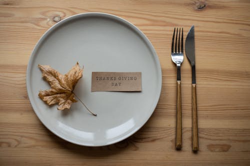 Top view layout of white porcelain plate with dry maple leaf and carton postcard with Thanks Giving Day Inscription placed in wooden table near fork and knife