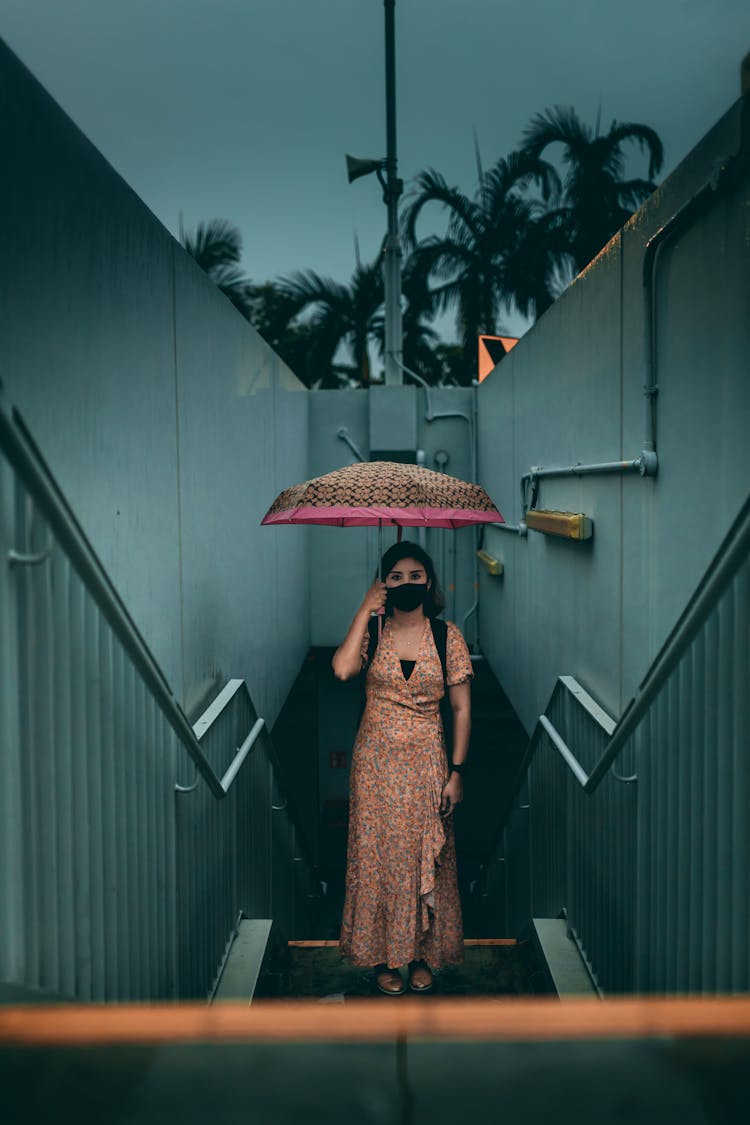 Woman In Face Mask Under Umbrella Standing On Street Steps