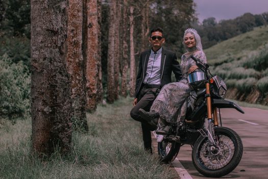 A fashionable couple posing with a motorcycle in a scenic forest setting, capturing happy expressions. - Fotografer Sidoarjo