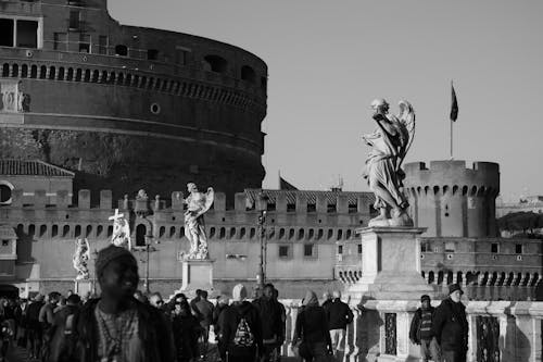 Black and white crowd of diverse travelers walking on bridge with marble statues near famous ancient Castel Sant Angelo on sunny day in Rome