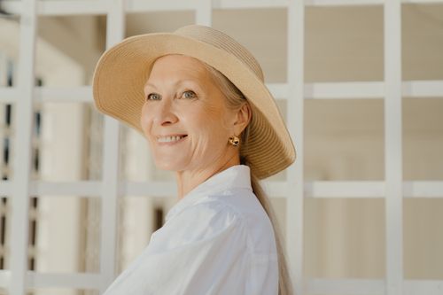 Free Senior woman smiling in an elegant sun hat and white blouse outdoors, radiating warmth. Stock Photo