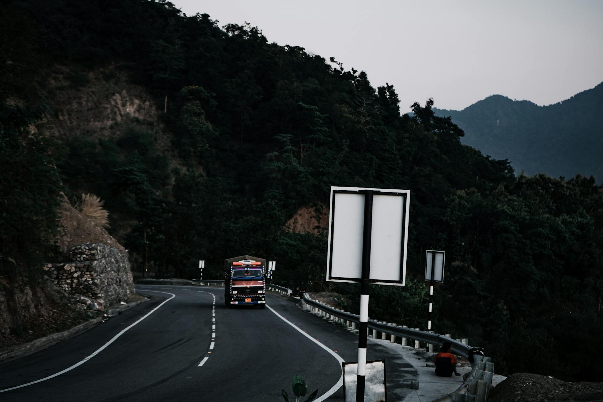 A truck navigates a winding mountain road surrounded by lush greenery and towering peaks.