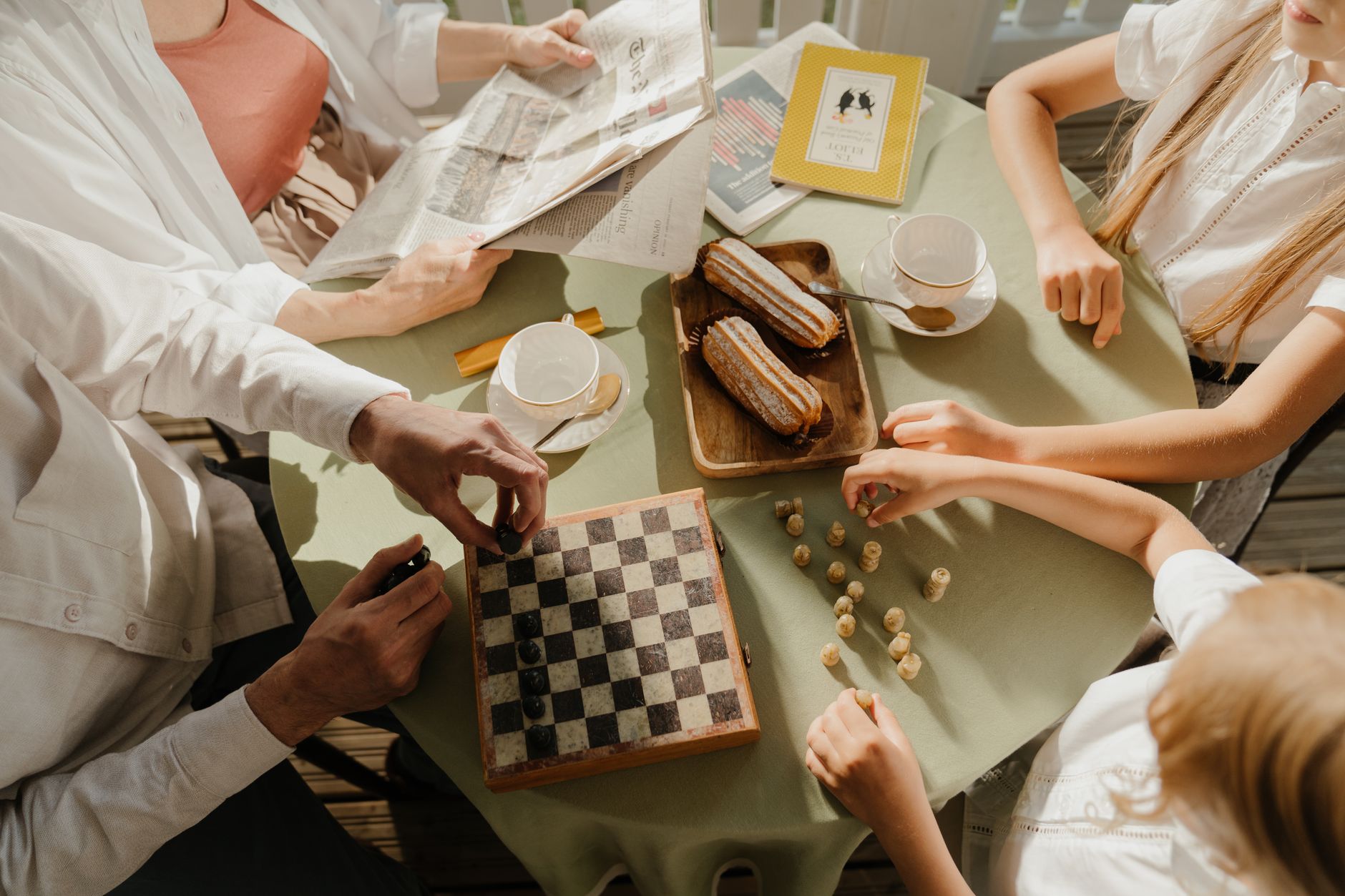 A family enjoys chess, tea, and pastries at a sunlit table, symbolizing togetherness.