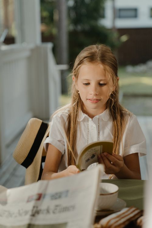 Free A young girl reads a book outdoors on a sunny porch, surrounded by a newspaper and a hat. Stock Photo