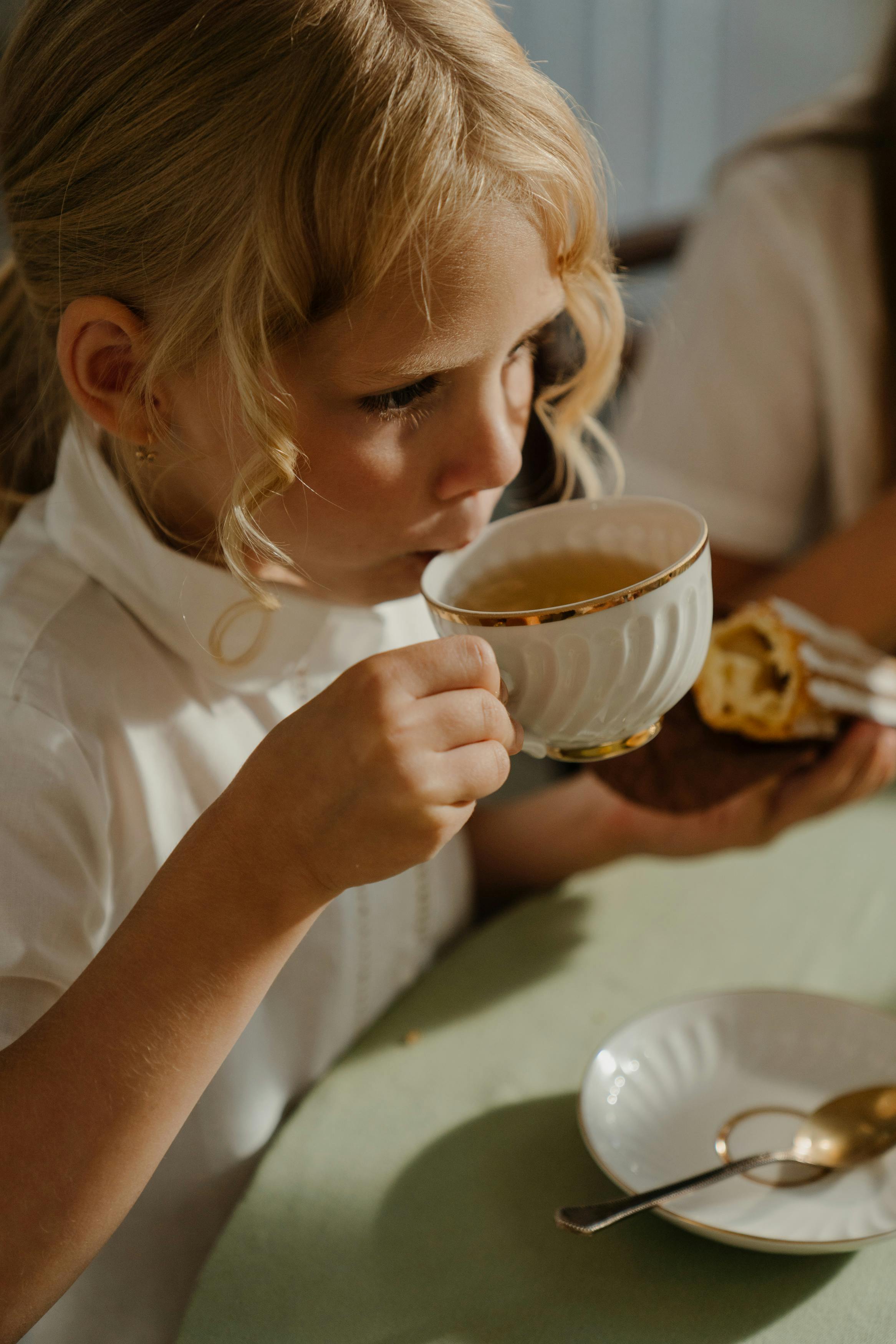 Charming child with blond hair sipping tea in a cozy indoor setting.