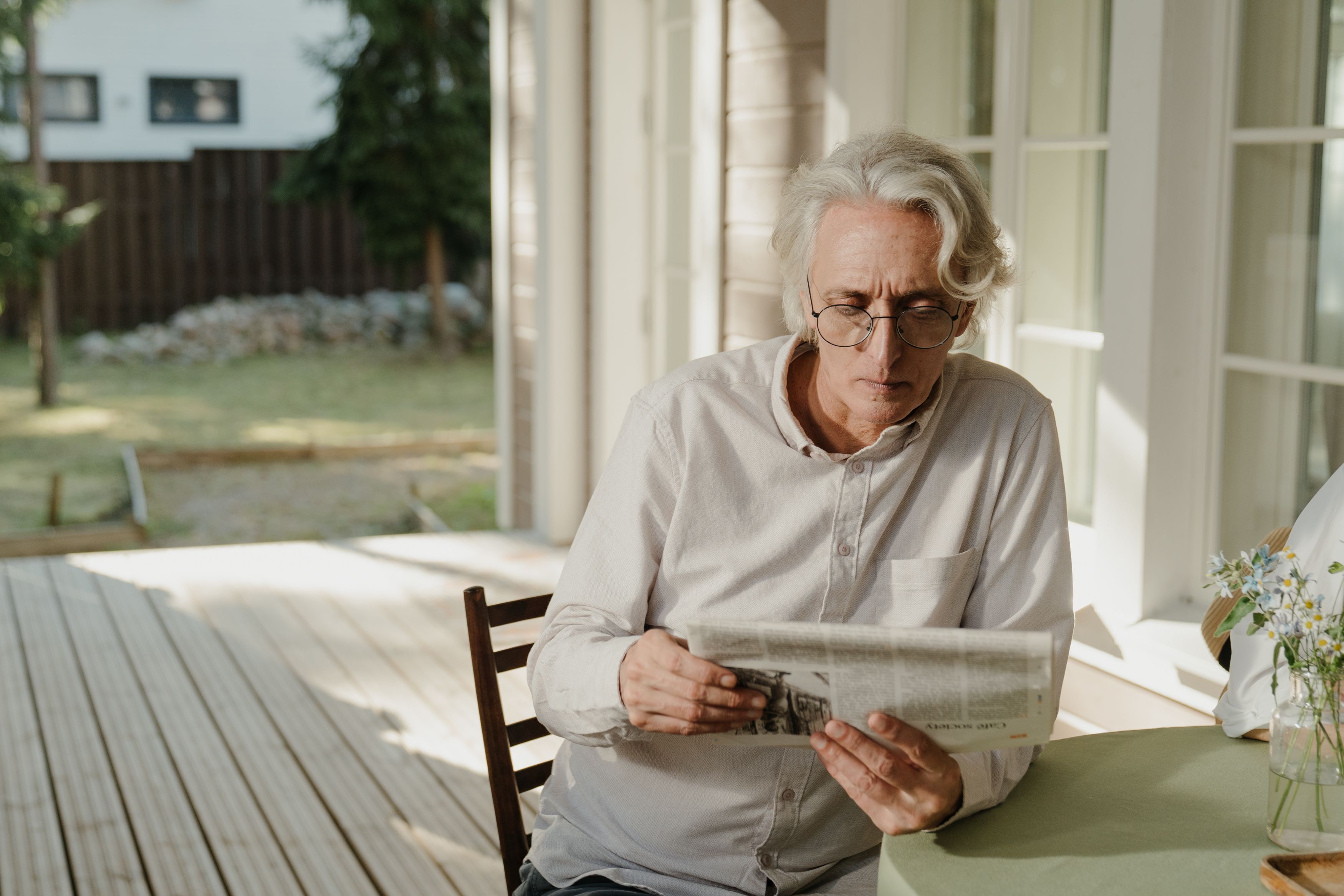 Elderly man with gray hair reading the newspaper on a sunny outdoor veranda.
