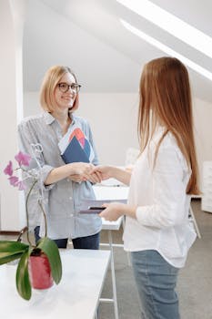Two women exchanging documents and handshaking in a modern office setting.