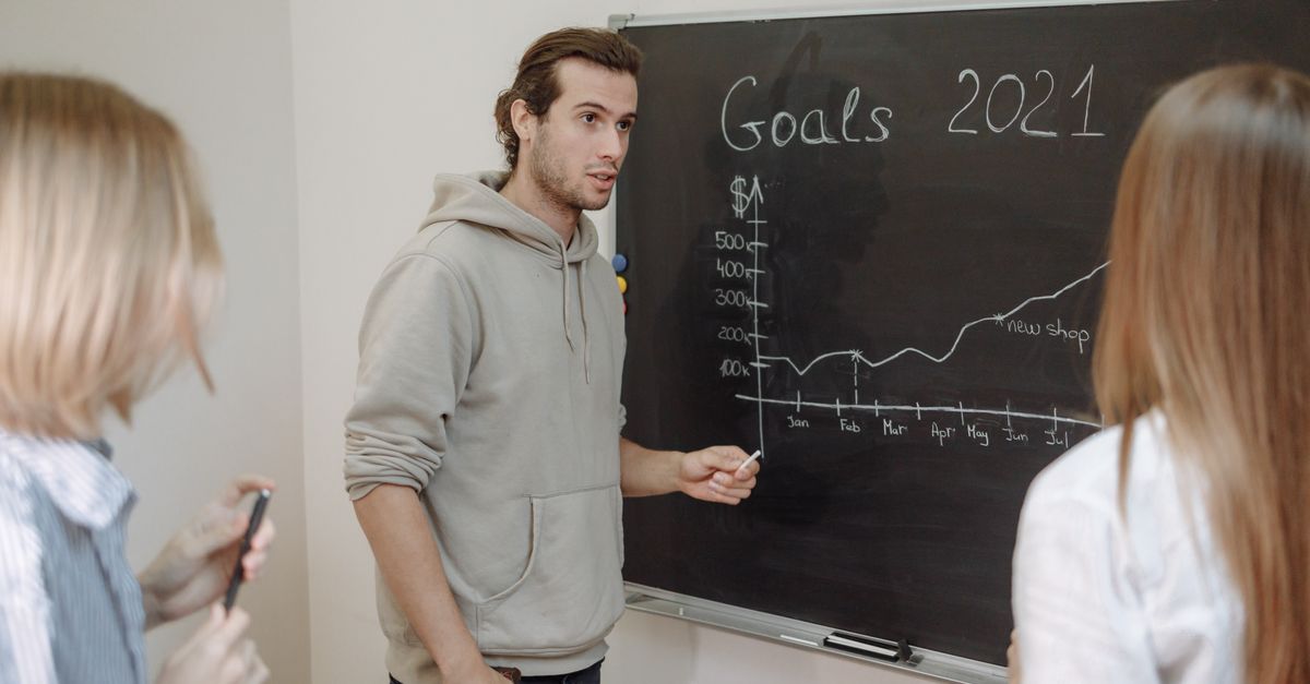 Three professionals engaged in a business discussion about goals written on a chalkboard in an office.