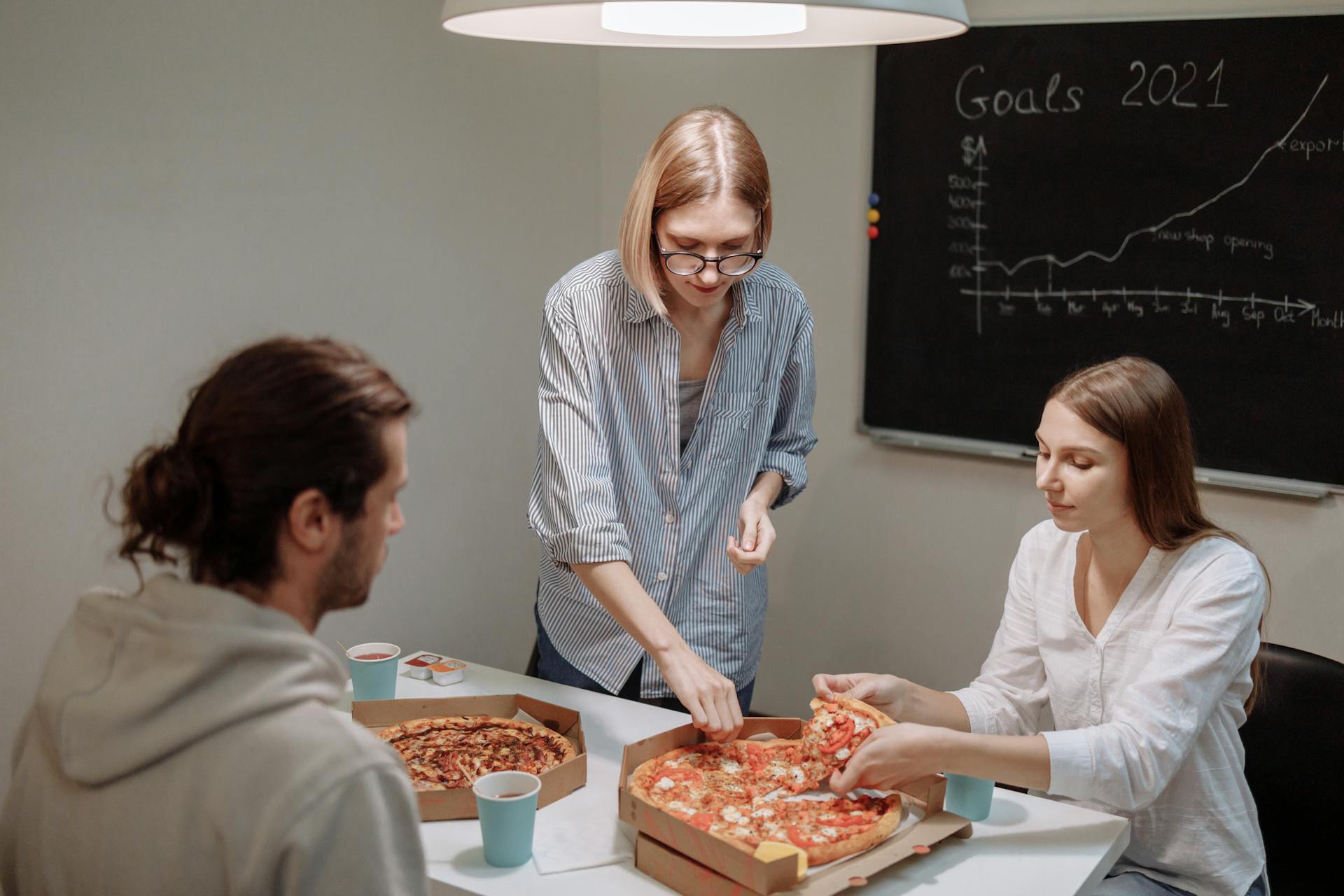 Office colleagues sharing pizza during a casual meeting with a goal board in the background.