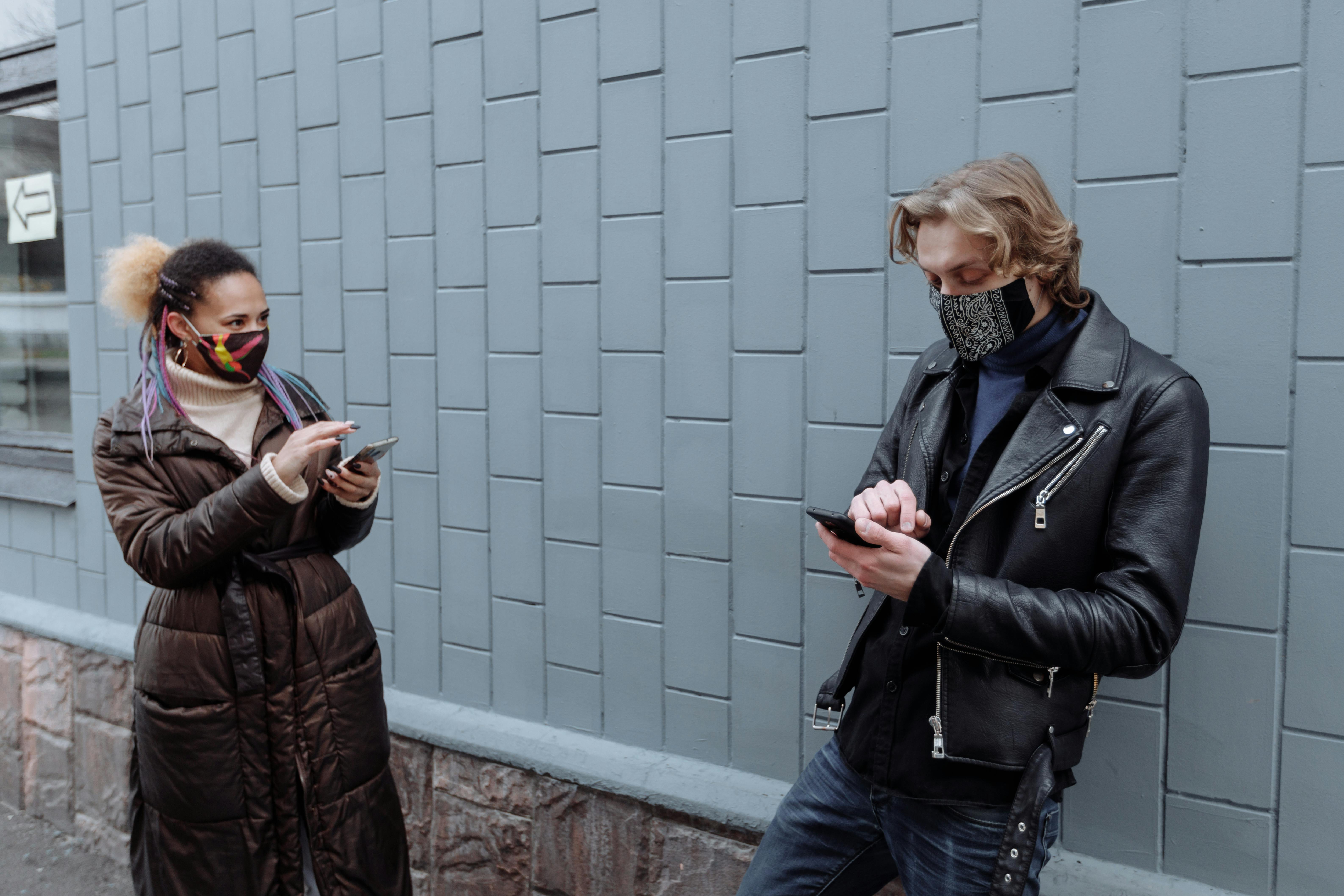 woman and a man wearing jackets and face masks using smartphones