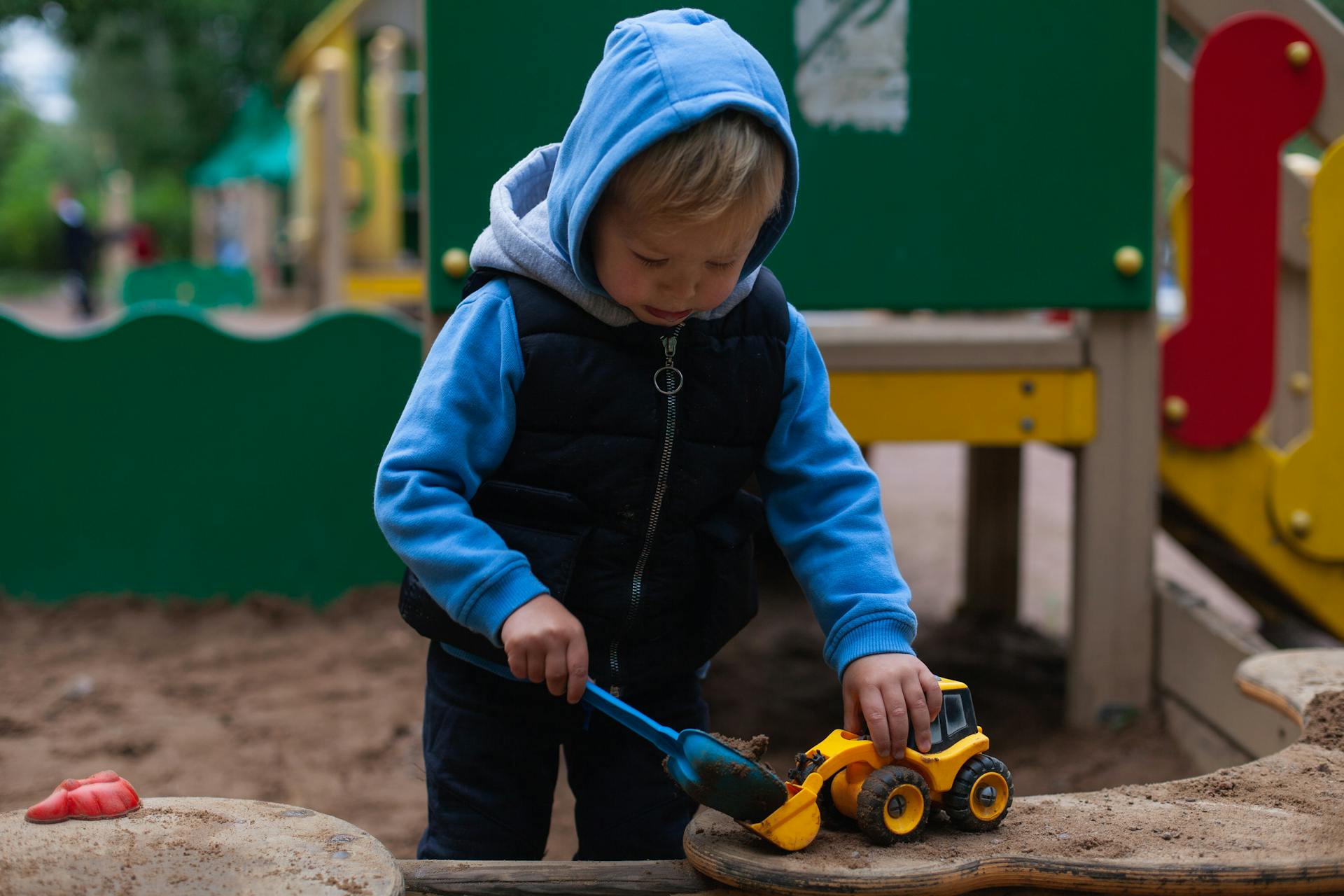 A young child plays with toy vehicles in a colorful outdoor playground sandbox.