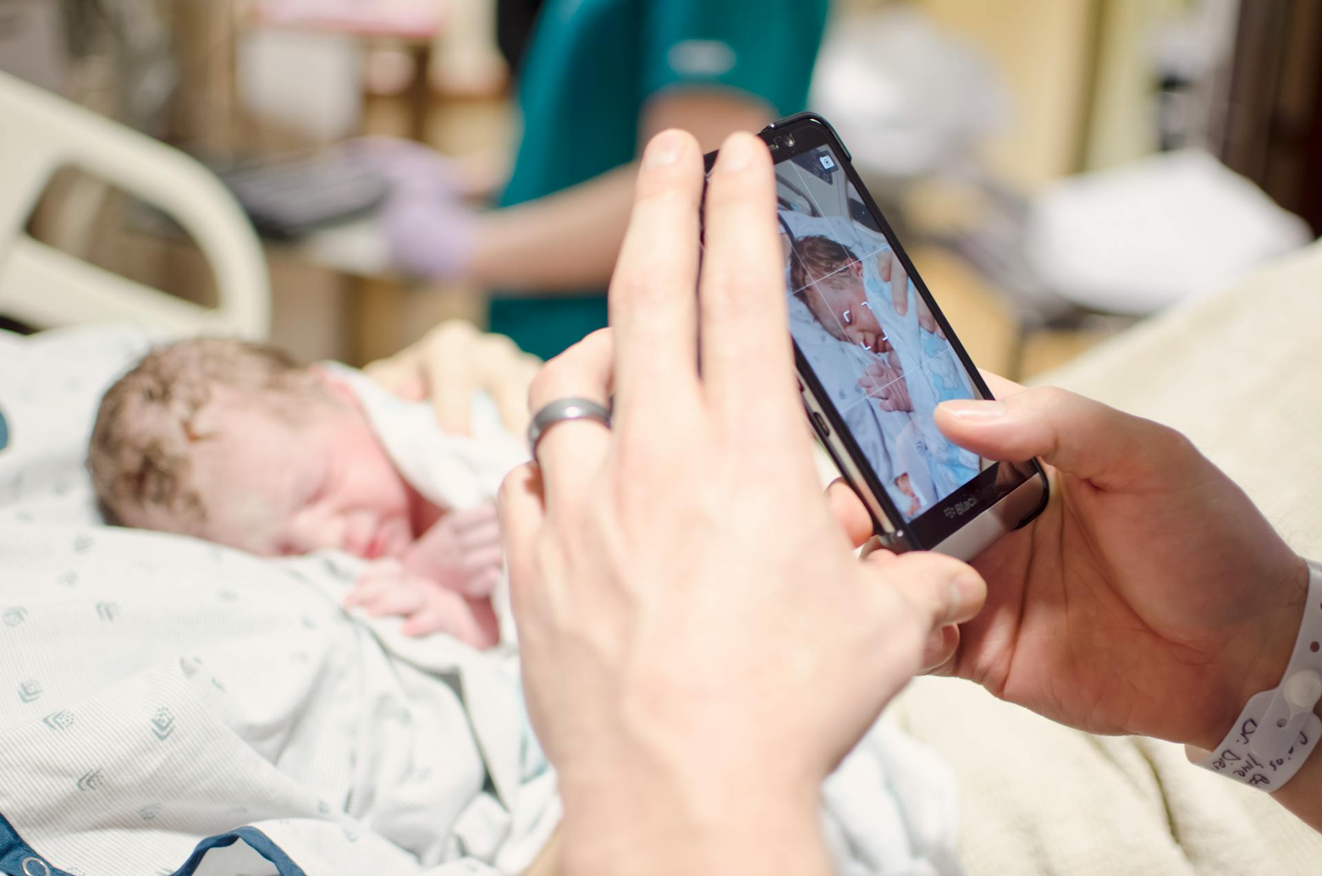 A close-up photo of a newborn baby being photographed with a smartphone in a hospital setting.