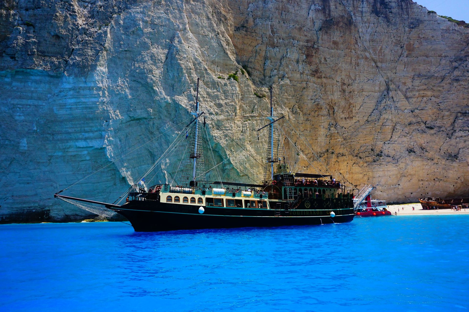 A historic ship by the rocky cliffs and vivid blue waters of Greece's coast.