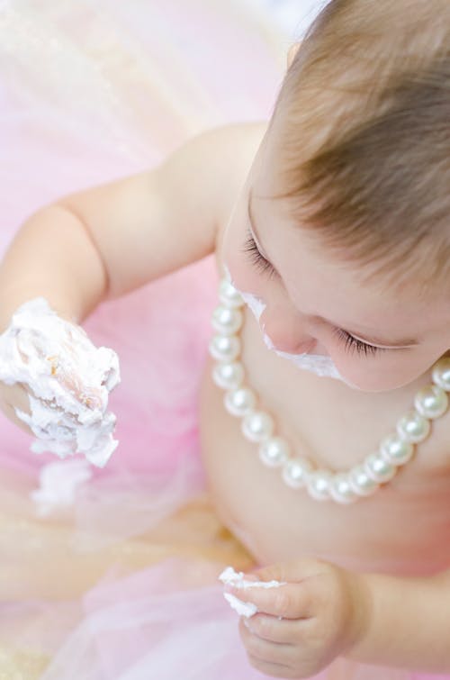 High-Angle Shot of a Baby Wearing a Pearl Necklace while Eating Frosting