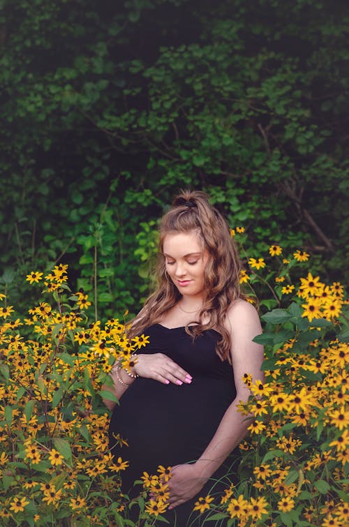 Photo of a Mother in a Black Dress Looking at Her Baby Bump Near Yellow Sunflowers