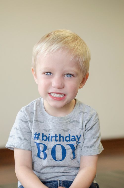 Free Young boy with blonde hair and blue eyes wearing a birthday shirt smiles indoors. Stock Photo