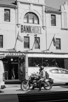Black and white photo of a biker riding by a historic hotel in a bustling city setting.