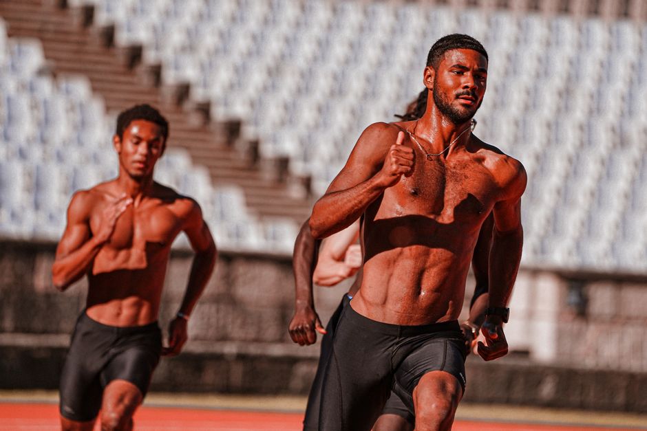 Shirtless male athletes displaying determination while sprinting on a sunny outdoor track.