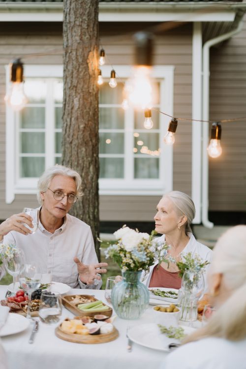 Free Senior adults dining outdoors, engaging in conversation at a beautifully set table with decor and string lights. Stock Photo