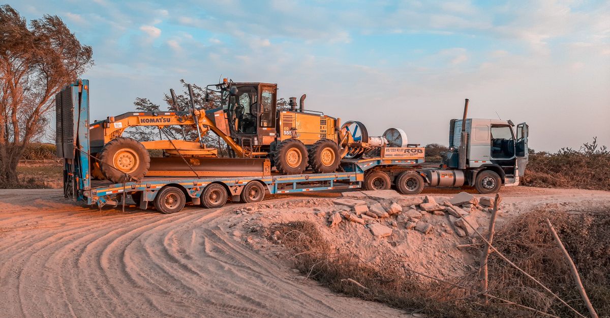 A cargo truck transporting heavy machinery on a rugged dirt road under a clear sky.