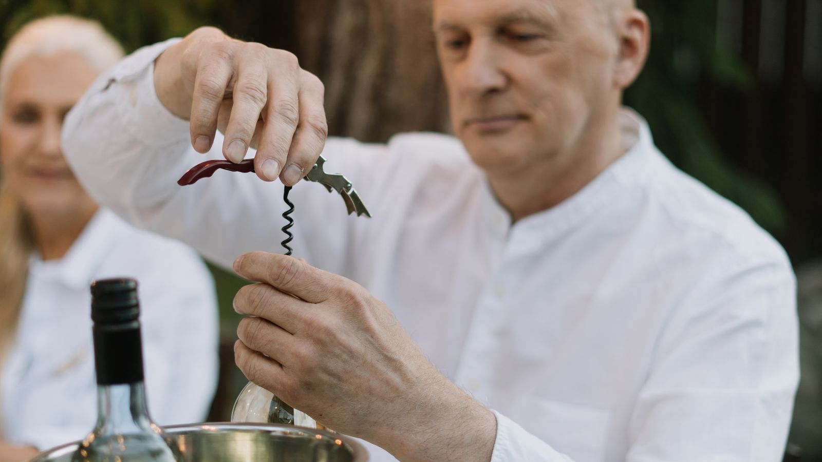 A man opening a bottle of wine with a corkscrew at an outdoor dinner table