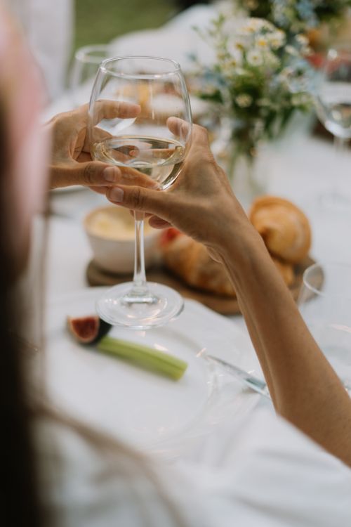 Free Close-up of hands holding a white wine glass at an elegant table setting with bread, cheese, and flowers. Stock Photo