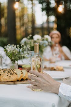 A hand holding a wine bottle in a scenic outdoor dinner setting with bread and flowers.
