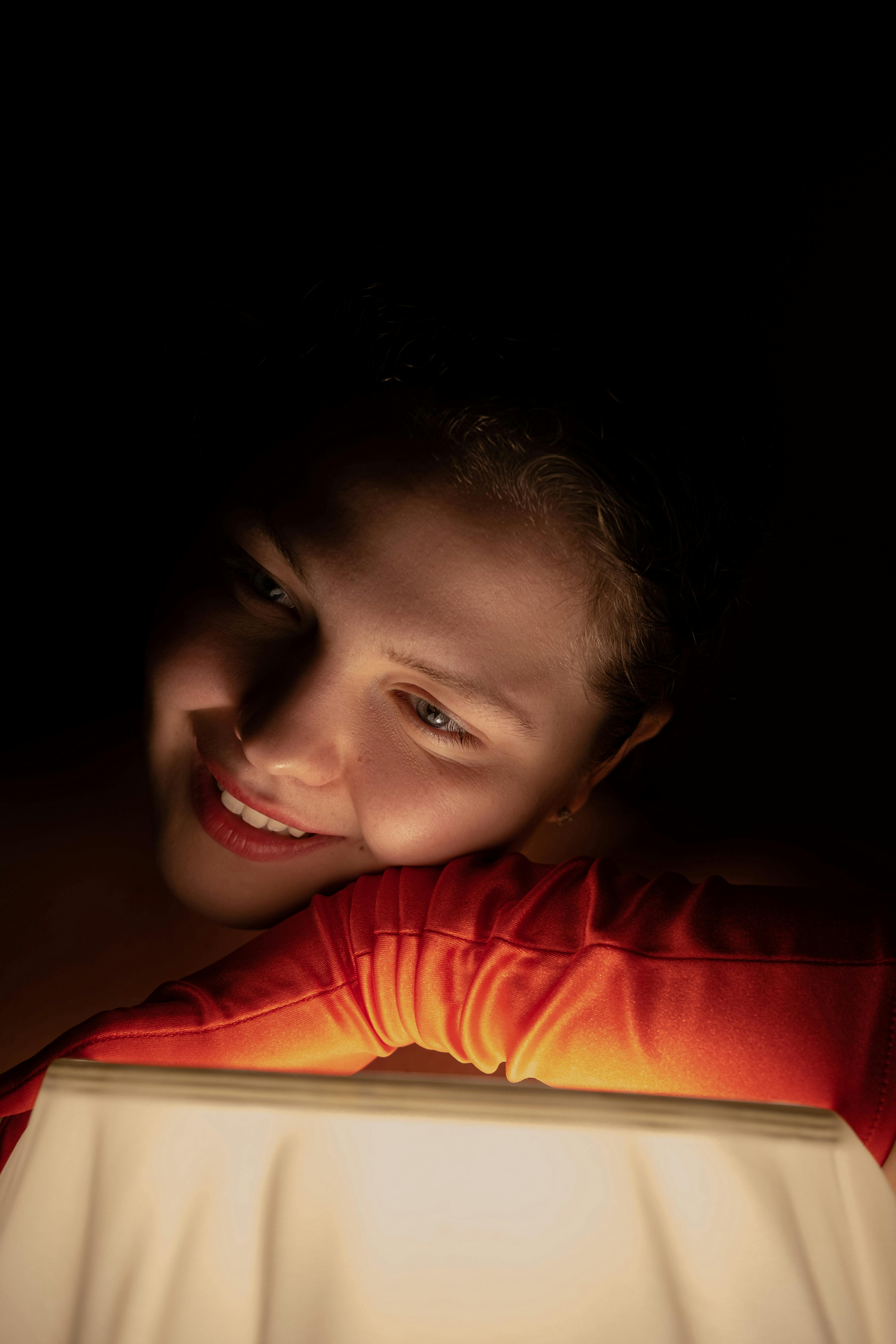 Happy mysterious female wearing red glove smiling and leaning on illuminated lamp while standing in dark room on night time