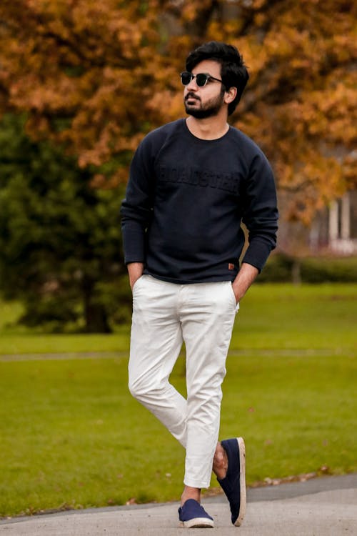 Man in Black Sweatshirt and White Trousers with Hands in Pockets Standing on One Leg in a Park