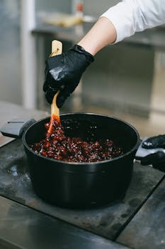 Chef wearing black gloves stirs a red fruit sauce in a black pot, creating a savory dish.