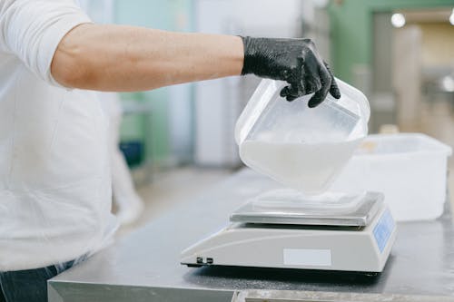 Person Pouring an Ingredient from a Plastic Container to a Weighing Scale