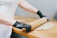 Close-Up Photo of a Person Wearing Black Gloves Flattening a Dough with a Rolling Pin