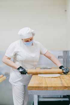 A baker dressed in white uniform and mask rolling dough on a wooden table.