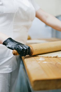 A pastry chef in black gloves rolling dough with a wooden pin in a kitchen.