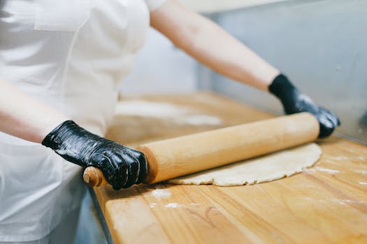 Pastry chef wearing gloves rolls out dough on a wooden table in a kitchen.