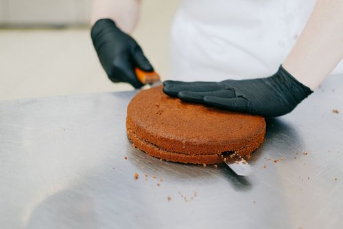 Free A baker in gloves skillfully slices a chocolate cake with a spatula on a metal table. Stock Photo
