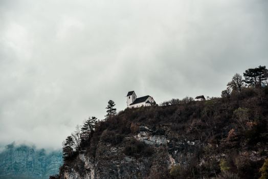 From below of rocky rough cliff covered with houses among dry plants under cloudy hazy sky